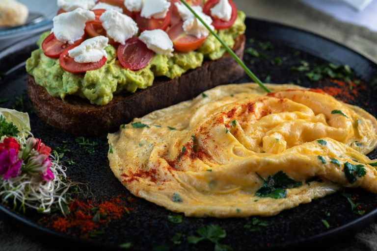 Vibrant avocado toast with cherry tomatoes and creamy omelette on a dark plate.