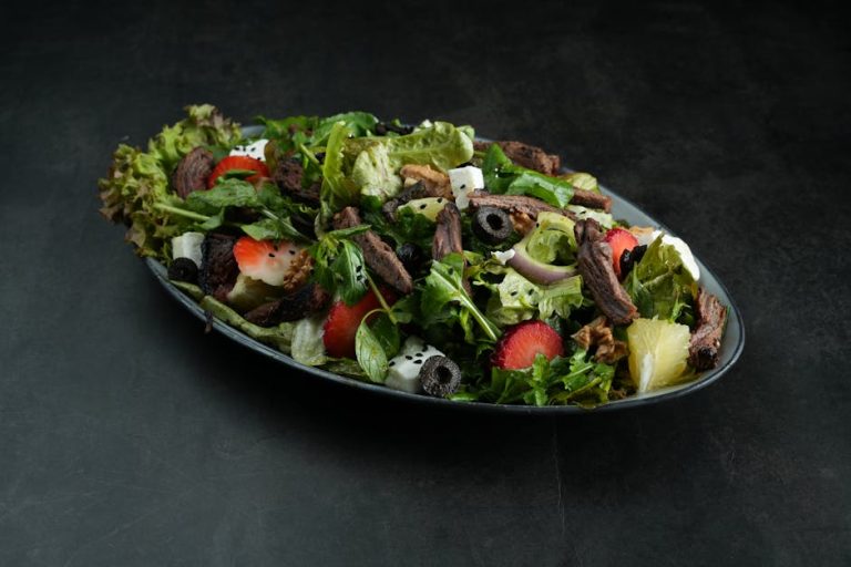 Vibrant mixed greens salad featuring fruit slices, grilled meat, and black olives on a dark background.