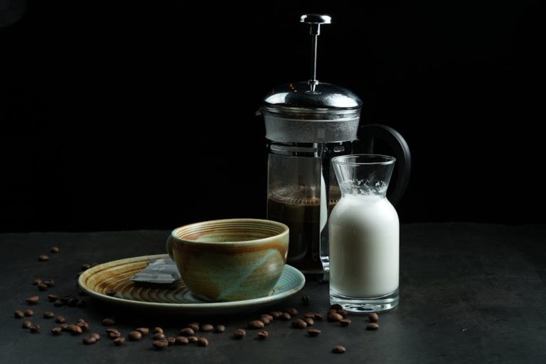Artistic display of a coffee setup featuring a French press, ceramic cup, milk jug, and coffee beans.