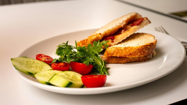 A close-up of a sandwich with tomatoes, parsley, and cucumbers on a white plate.