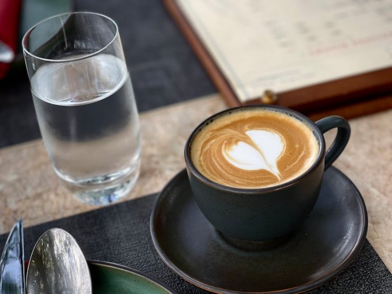 Close-up of a latte with heart-shaped latte art beside a glass of water on a cafe table.