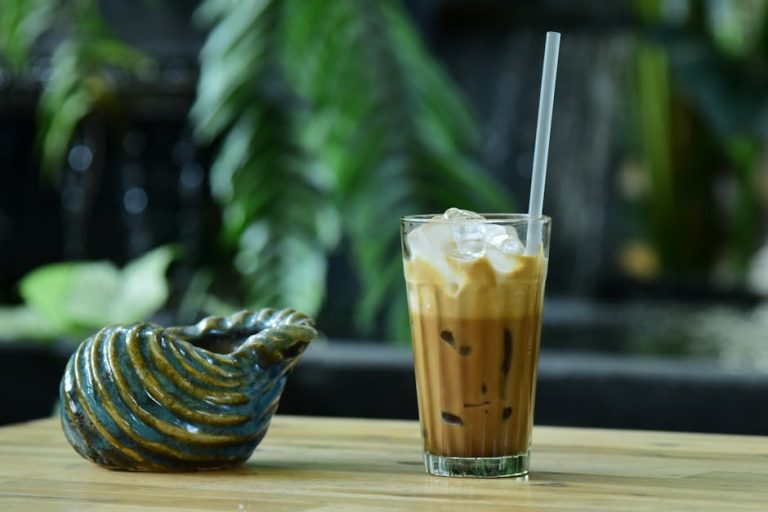 Refreshing iced latte with straw beside artistic ceramic vase on a wooden table outdoors.