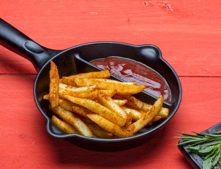 Crispy seasoned French fries with ketchup in a skillet on a red wooden table.