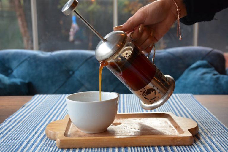 Hand pouring fresh coffee from a French press into a ceramic cup on a wooden tray indoors.
