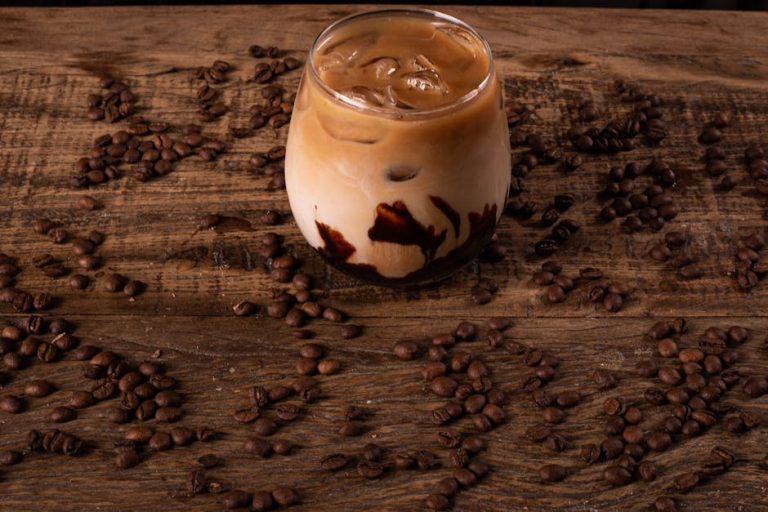 A cold iced coffee with chocolate syrup surrounded by coffee beans on a rustic wooden table.